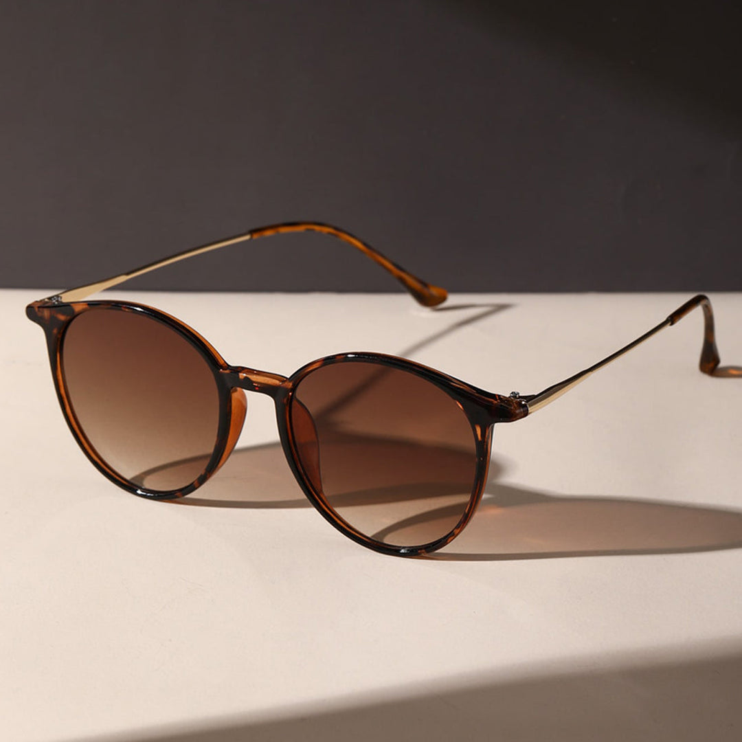 Round tortoiseshell sunglasses with brown gradient, anti-glare lenses are placed on a light-colored surface, casting a shadow. The dark background highlights the eyewears stylish UV protection.