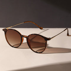 Round tortoiseshell sunglasses with brown gradient, anti-glare lenses are placed on a light-colored surface, casting a shadow. The dark background highlights the eyewears stylish UV protection.
