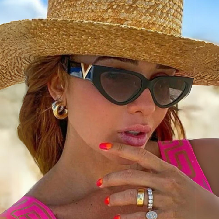 A woman in a wide-brimmed straw hat and vintage cat eye sunglasses poses with her hand near her lips, showing off bright orange nails and multiple rings. She has gold hoop earrings and wears a pink top.