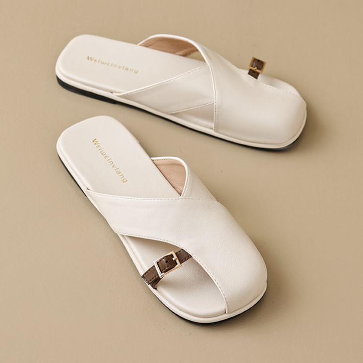 A pair of chic slides, featuring white crisscross straps, brown buckle details, and flat black soles, are displayed on a beige surface—perfect for elevating your summer wardrobe.
