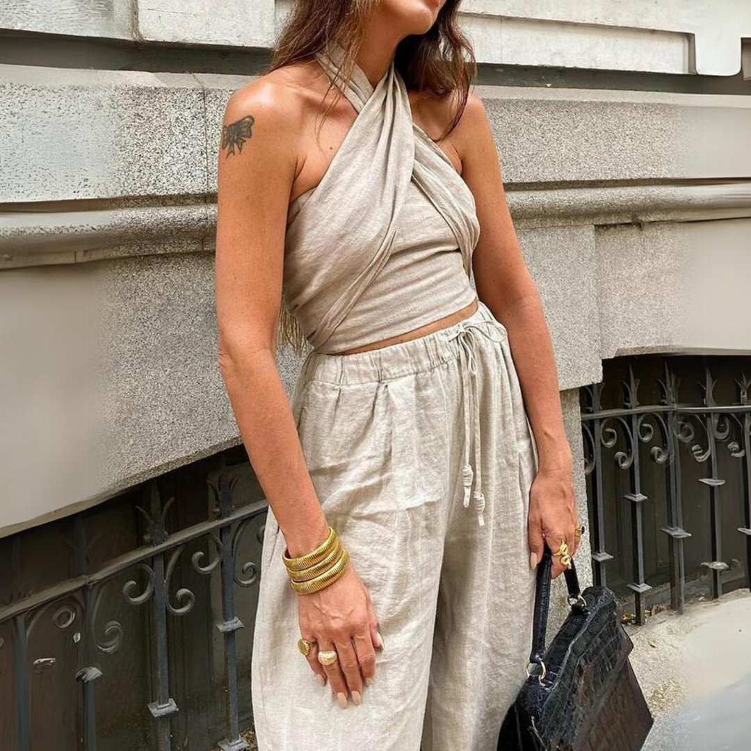 Woman standing outdoors in a light beige wrap crop top and chic halter collar, paired with loose wide-leg pants, accessorized with gold bangles, rings, a black textured handbag, and a visible tattoo on her upper arm.