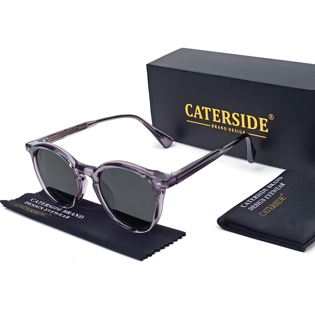 Translucent gray round frame sunglasses with black lenses are displayed next to a black Caterside box, a branded cleaning cloth, and a soft pouch—perfect for those seeking fashionable eyewear on a white background.