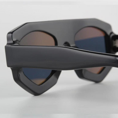 Close-up of black acetate geometric sunglasses with thick frames and dark, slightly reflective lenses, offering UV400 protection while resting on a white surface.
