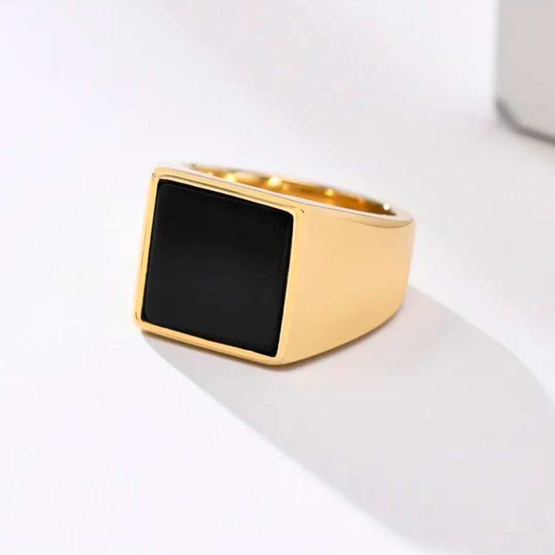 A gold ring with a thick band and a large, square black stone centerpiece is displayed on a white surface under soft lighting, showcasing its polished finish for versatile styling.