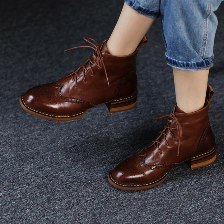 A person wearing rolled-up blue jeans and genuine leather brogue boots stands on a dark carpeted floor, capturing the essence of an urban trendsetter.
