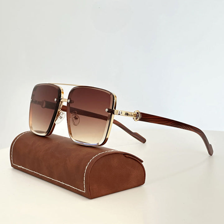 A pair of large, vintage sunglasses with gold frames, brown gradient lenses, and wooden arms rests on a brown, suede-textured case against a plain white background.