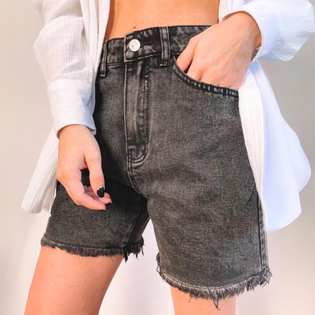 A person wearing high-waisted, black wash denim shorts with frayed hems and a white, partially open long-sleeve shirt stands with one hand in their pocket against a plain background.