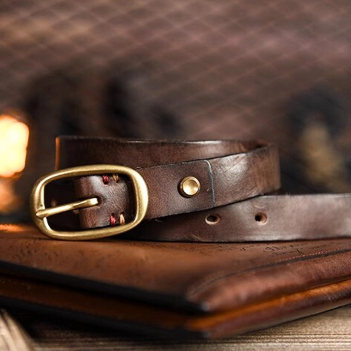A close-up of a brown leather belt with a brass buckle rests on top of a leather-bound book, capturing the timeless charm of country fashion against a softly blurred, warm-toned background.