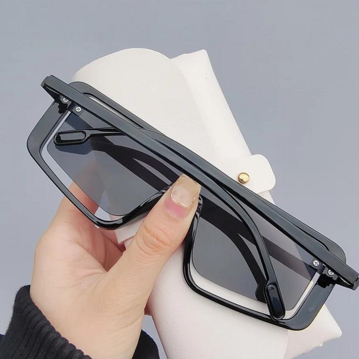 A hand holding black rectangular sunglasses with UV400 lens and gradient lenses, positioned in front of a white, textured eyeglasses case against a plain grey background.
