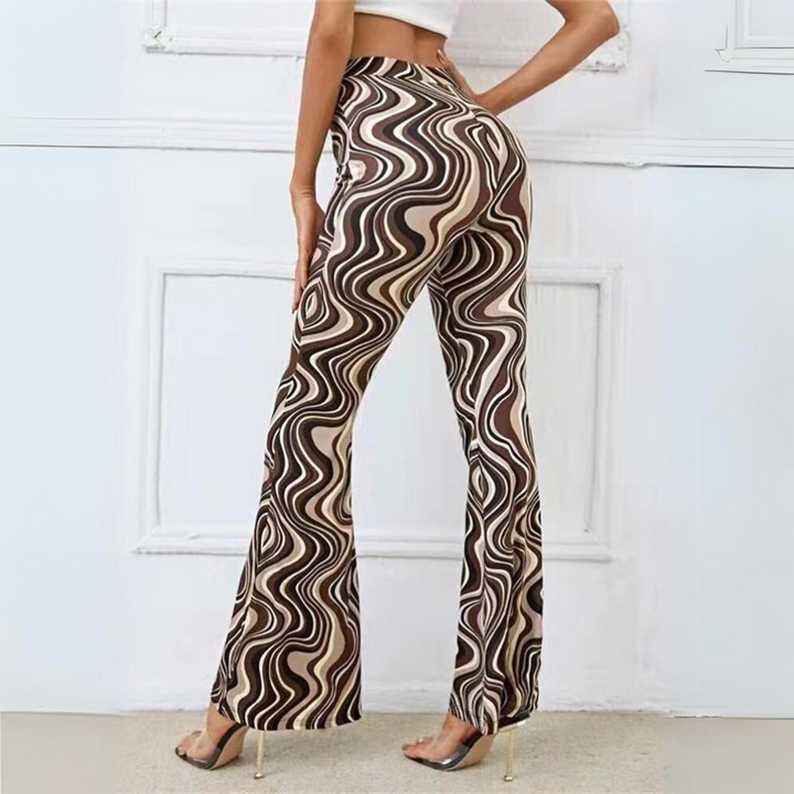 A person stands with their back to the camera, wearing trendy statement pants featuring a brown, beige, and black wavy swirl pattern and a white crop top, posing in high-heeled clear shoes against a white wall.