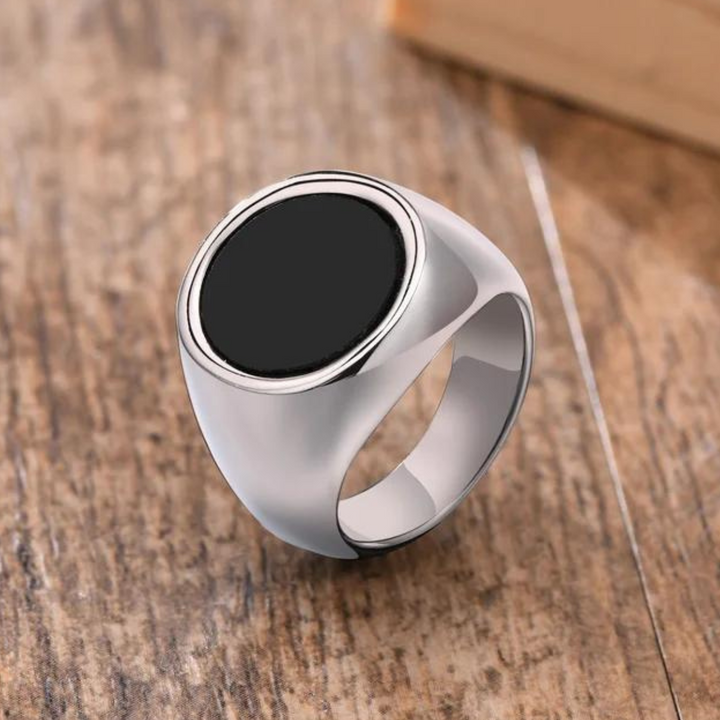 A silver signet ring with a large, flat black circular stone and a polished finish is displayed on a wooden surface, perfect for versatile styling.