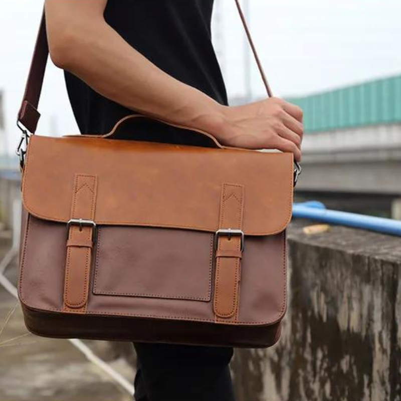 A person in a black shirt holds a brown premium leather messenger bag with an adjustable shoulder strap and front buckles, standing outdoors near a concrete wall.