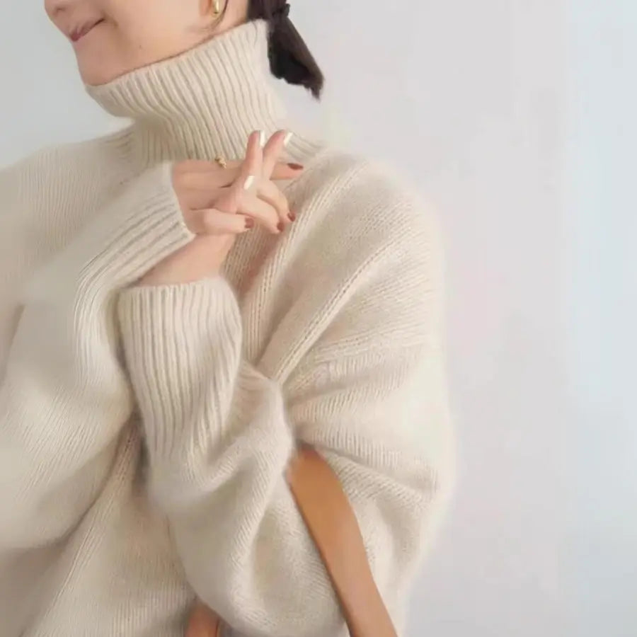 A person wearing a cozy cream-colored cashmere turtleneck sweater smiles softly, with hands gently clasped near the face and a brown strap resting on their shoulder. The background is plain and light.
