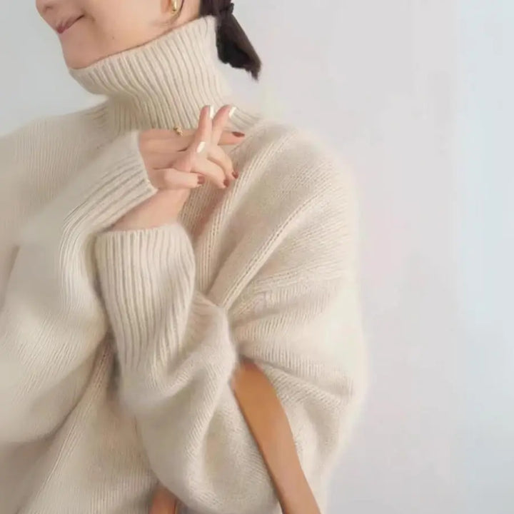 A person wearing a cozy cream-colored cashmere turtleneck sweater smiles softly, with hands gently clasped near the face and a brown strap resting on their shoulder. The background is plain and light.