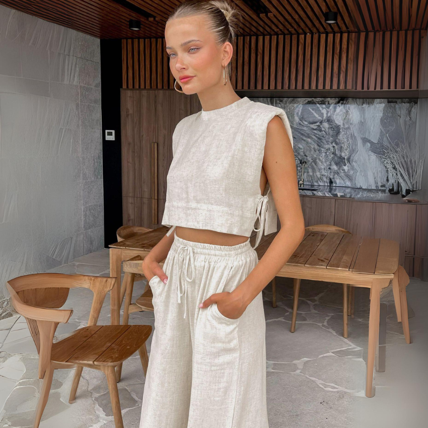 A woman in a light beige Belgian Linen Blend cropped top and matching wide-leg pants stands indoors with her hands in her pockets, surrounded by modern wooden furniture and a marble wall.