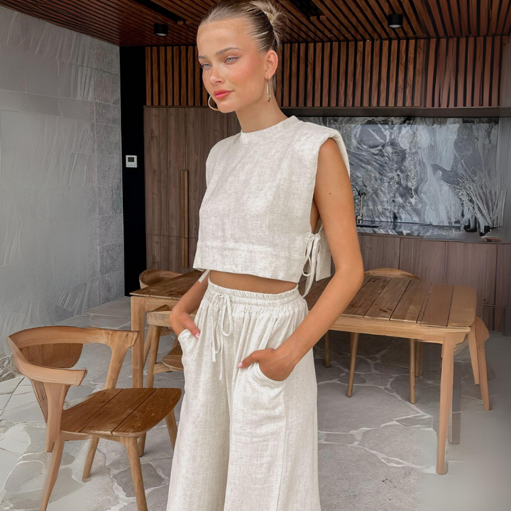 A woman in a light beige Belgian Linen Blend cropped top and matching wide-leg pants stands indoors with her hands in her pockets, surrounded by modern wooden furniture and a marble wall.