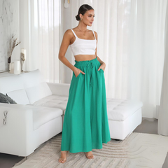 A woman stands indoors near a white sofa, wearing a white crop top and a long, high-waisted teal-green skirt with an elastic waistband and relaxed fit. The room is bright with sheer curtains and neutral decor.