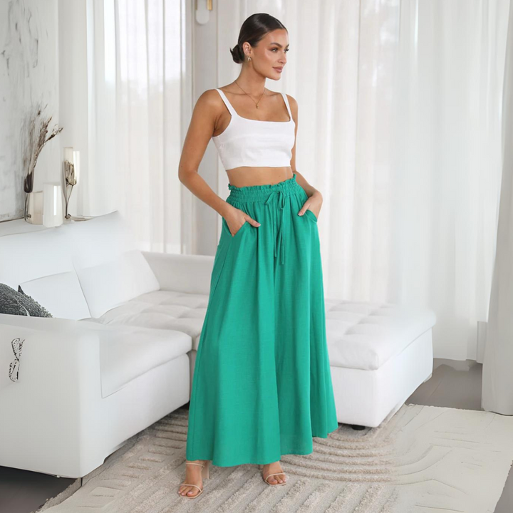 A woman stands indoors near a white sofa, wearing a white crop top and a long, high-waisted teal-green skirt with an elastic waistband and relaxed fit. The room is bright with sheer curtains and neutral decor.