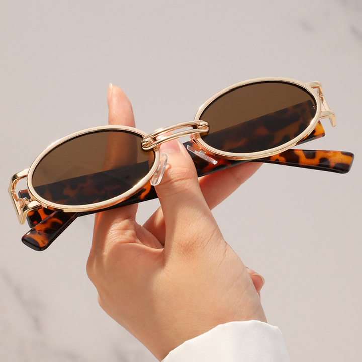 A hand holds retro oval sunglasses with brown lenses and a durable metal frame. The gold temples feature a brown tortoiseshell pattern, while the softly blurred, light background highlights their stylish UV400 protection.