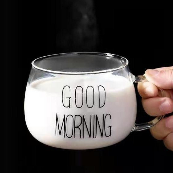 A hand holds a clear glass mug with an ergonomic handle, filled with milk. The mug has the words GOOD MORNING printed in bold, black letters. The black background highlights its modern design.