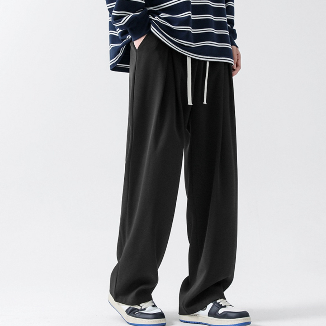 A person wearing loose black streetwear joggers with a white drawstring, a navy and white striped long-sleeve shirt, and blue-and-white sneakers, standing against a plain light background.