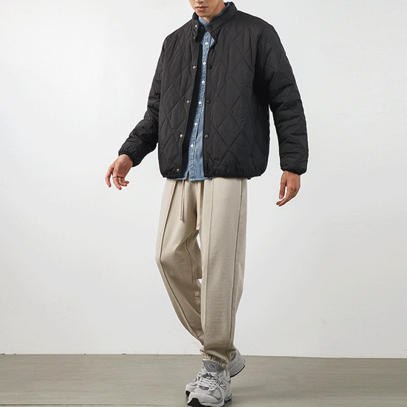 A person stands against a plain background wearing a black diamond-stitched padded jacket over a blue button-up shirt, beige loose-fitting trousers, and gray sneakers. Their face is partially out of frame.