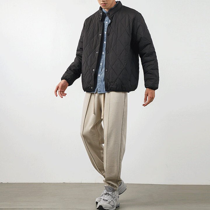 A person stands against a plain background wearing a black diamond-stitched padded jacket over a blue button-up shirt, beige loose-fitting trousers, and gray sneakers. Their face is partially out of frame.