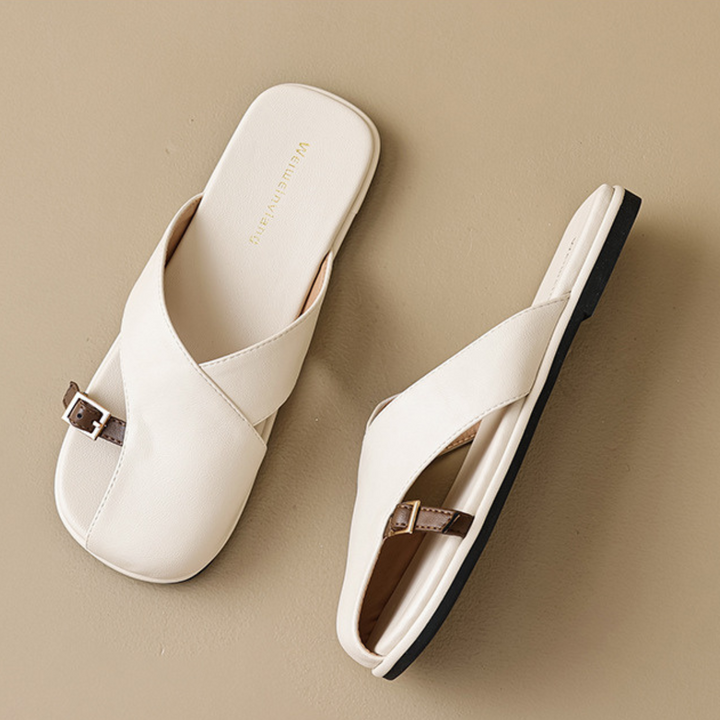 A pair of chic off-white leather slides with crisscross straps and small brown buckles, perfect for elevating your summer wardrobe, displayed on a light beige surface.