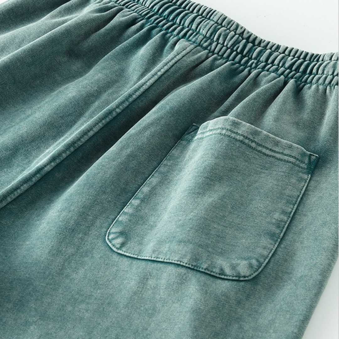 Close-up of greenish-blue elastic waistband shorts featuring a single back pocket on one side. The fabric appears soft and slightly faded, suggesting a casual, comfortable style with an easy elastic waistband for all-day wear.