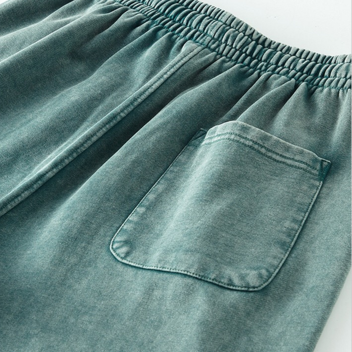 Close-up of greenish-blue elastic waistband shorts featuring a single back pocket on one side. The fabric appears soft and slightly faded, suggesting a casual, comfortable style with an easy elastic waistband for all-day wear.