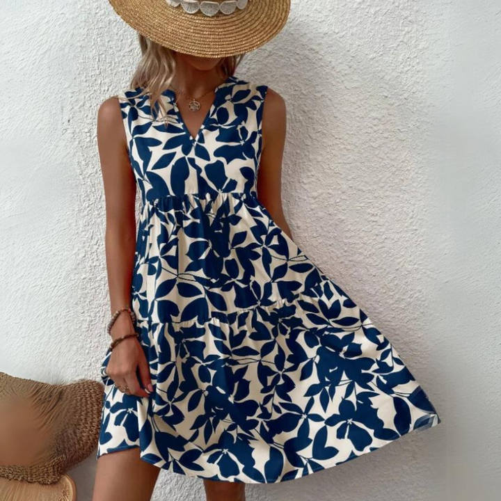 A person wearing a sleeveless, blue and white floral dress made from lightweight breathable fabric and a wide-brimmed straw hat stands against a textured white wall, holding the side of the dress to display its loose, flowing fit.