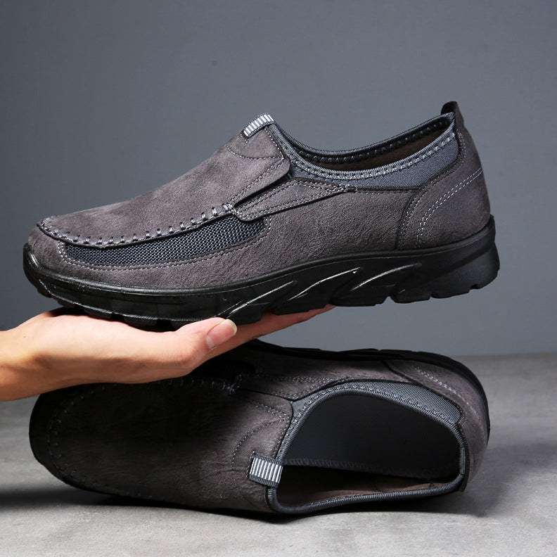 A pair of gray Men Leather Casual Shoes is displayed, with one shoe held above the other by a hand. The shoes feature a suede-like texture, black rubber soles, black mesh accents, and offer a comfortable fit.