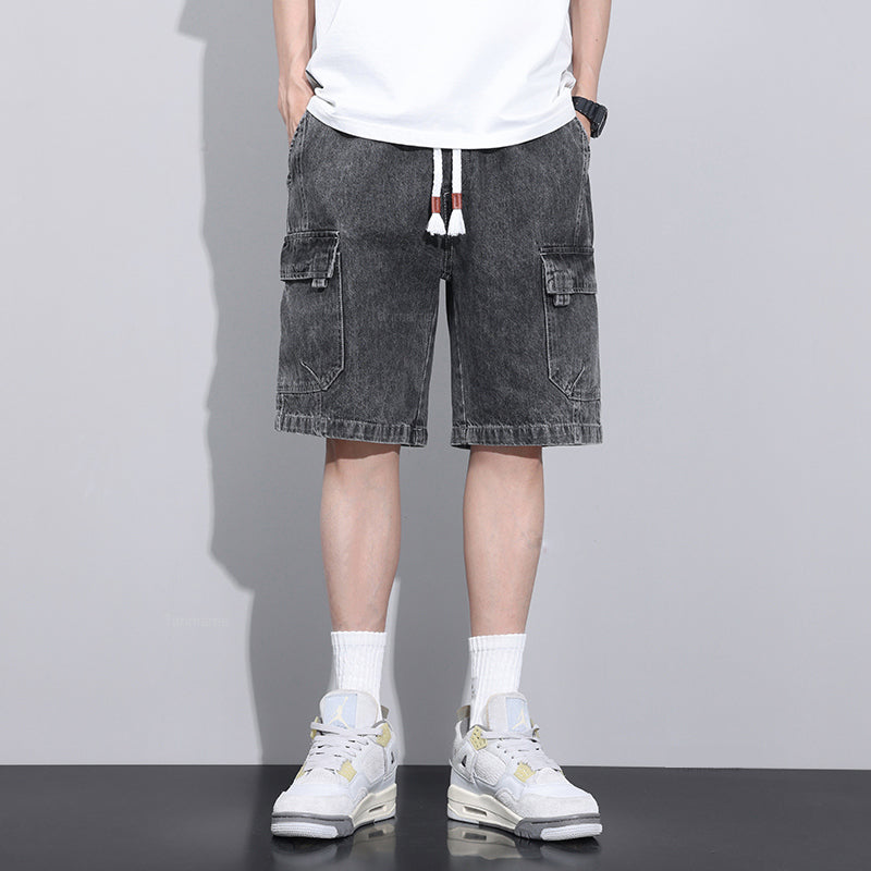 A person wearing a white t-shirt, mens denim shorts with an elastic waist and drawstrings, white crew socks, and white sneakers stands against a plain gray background. Their hands are in their pockets, and their face is not visible.