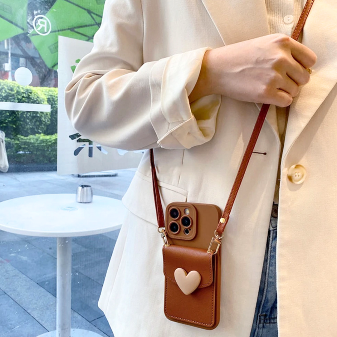 A person in a cream blazer holds a brown purse with a chic phone case and beige heart design, standing indoors near a window with greenery and outdoor seating visible outside.