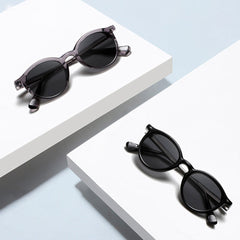Two pairs of fashionable eyewear—round frame sunglasses with transparent gray and solid black frames—are displayed on separate white rectangular surfaces against a light blue background.
