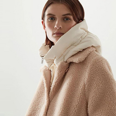 A person with straight brown hair wears a beige teddy coat and a large, padded off-white scarf made from durable material against a plain, light background.