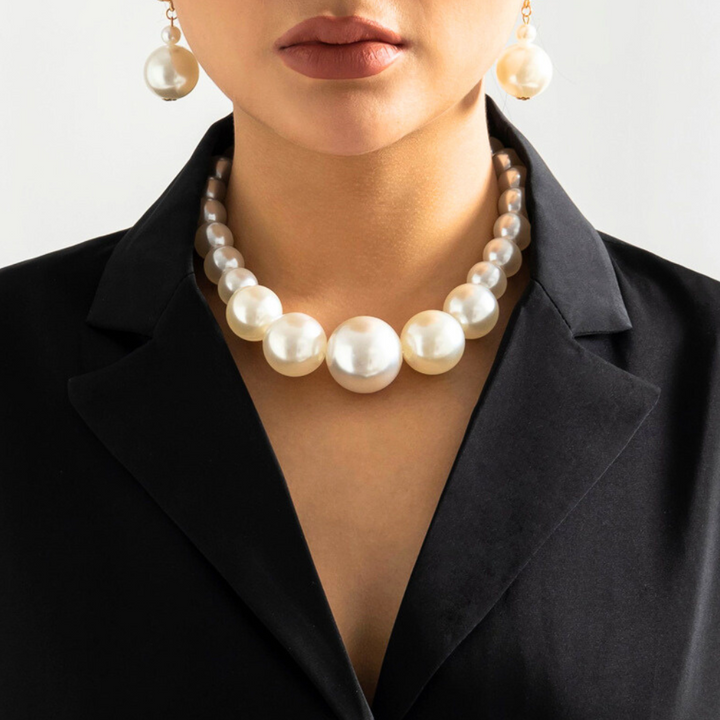 A woman wearing a black blazer and a statement pearl jewelry set—large necklace and matching earrings. The image is cropped to show her lower face, neck, and upper chest, highlighting the statement jewelry.