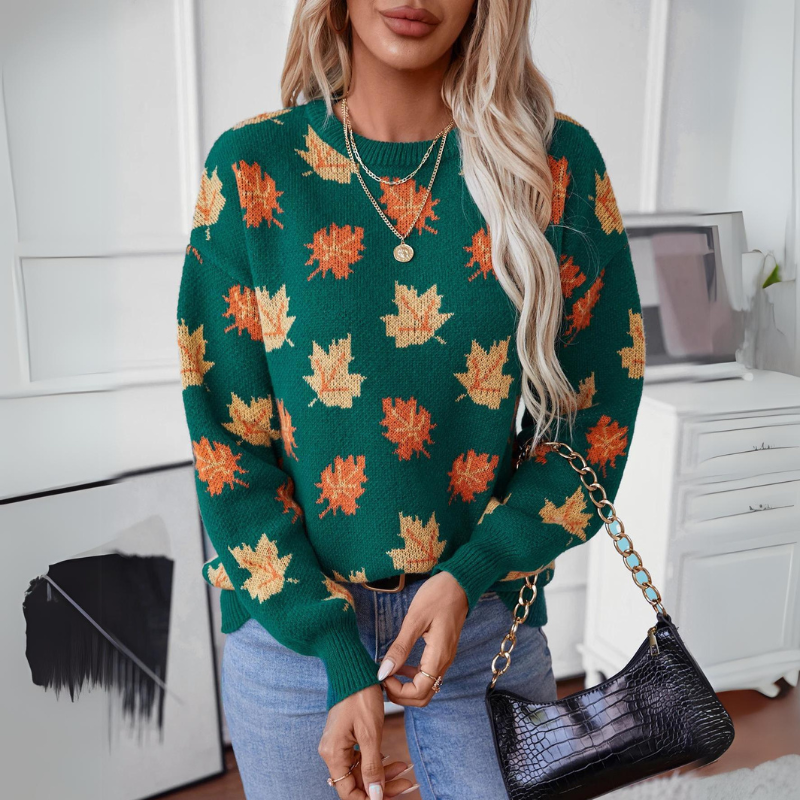 A woman with long blonde hair wears a comfortable green sweater featuring a maple leaf pattern, blue jeans, gold jewelry, and holds a black purse with a chain strap, standing in a modern, minimalistic room.