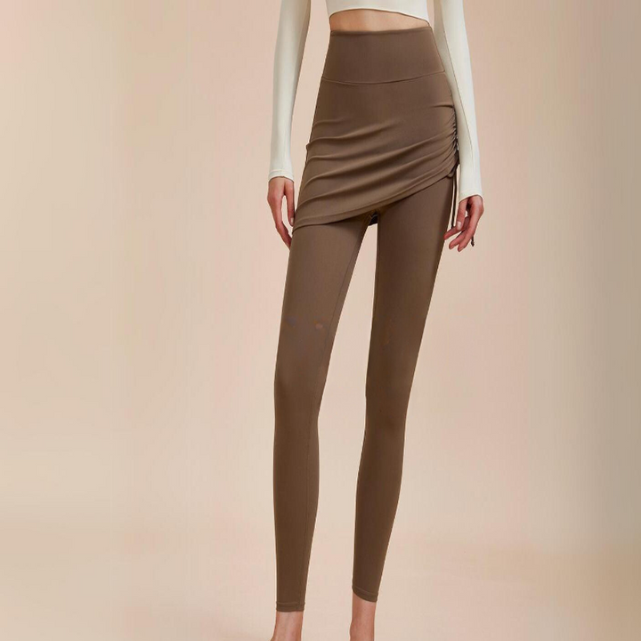 A person wearing high-waisted leggings with a matching asymmetrical skirt overlay and a long-sleeve white crop top, standing against a beige background—an ideal look for any autumn wardrobe.