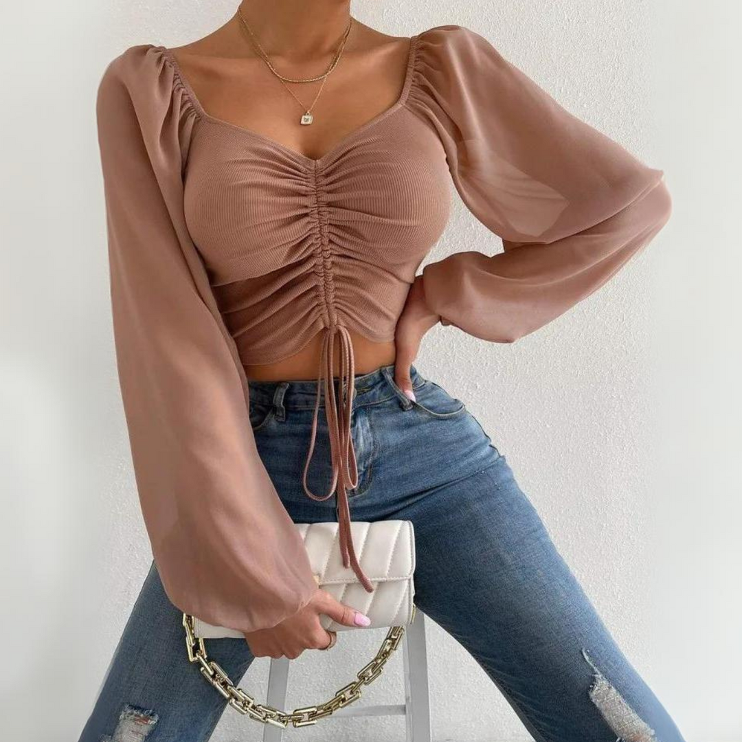 A person wearing a mauve crop top with puffed sleeves and a ruched front, blue ripped jeans, and holding a white purse with a gold chain, posing against a plain white background.