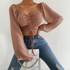 A person wearing a mauve crop top with puffed sleeves and a ruched front, blue ripped jeans, and holding a white purse with a gold chain, posing against a plain white background.