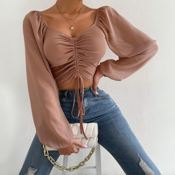 A person wearing a mauve crop top with puffed sleeves and a ruched front, blue ripped jeans, and holding a white purse with a gold chain, posing against a plain white background.