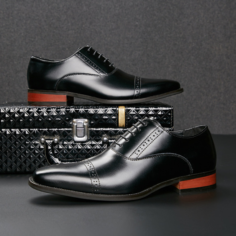 Two black Italian leather dress shoes with decorative broguing are displayed, one resting on a textured black briefcase and the other in front. Both shoes feature brown soles and are set against a dark background.