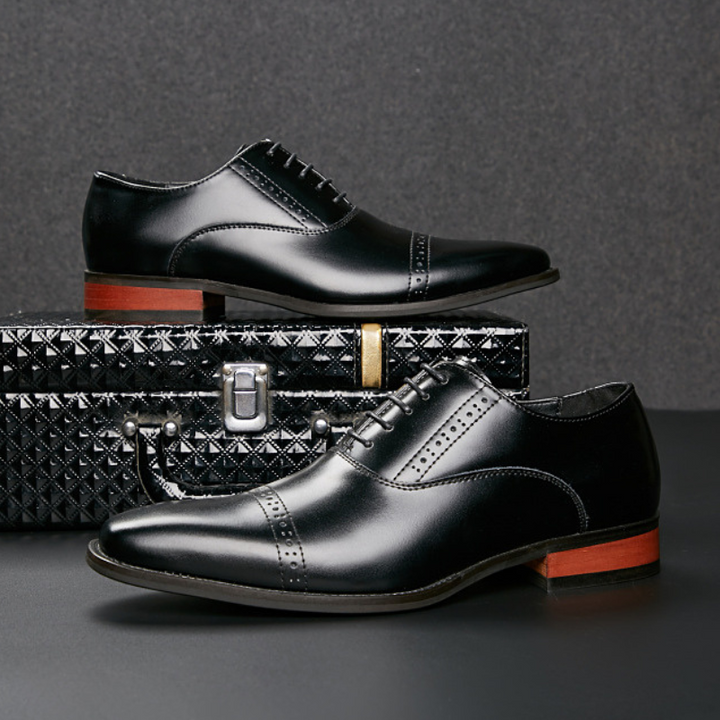 Two black Italian leather dress shoes with decorative broguing are displayed, one resting on a textured black briefcase and the other in front. Both shoes feature brown soles and are set against a dark background.