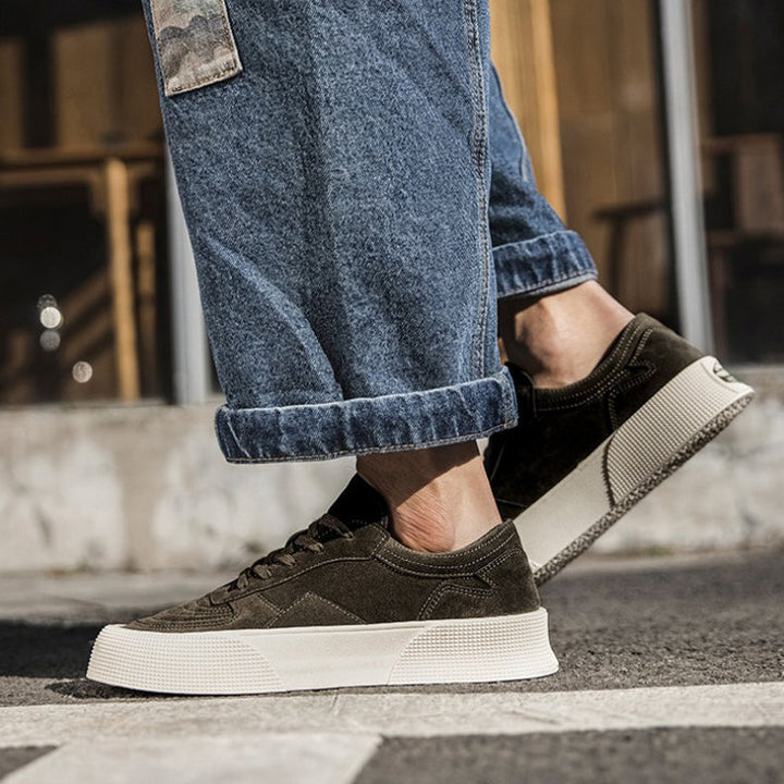 A person wearing loose-fitting blue jeans and dark green lace-up sneakers with comfortable cushioning and thick white soles stands on a street, with a blurred building in the background.