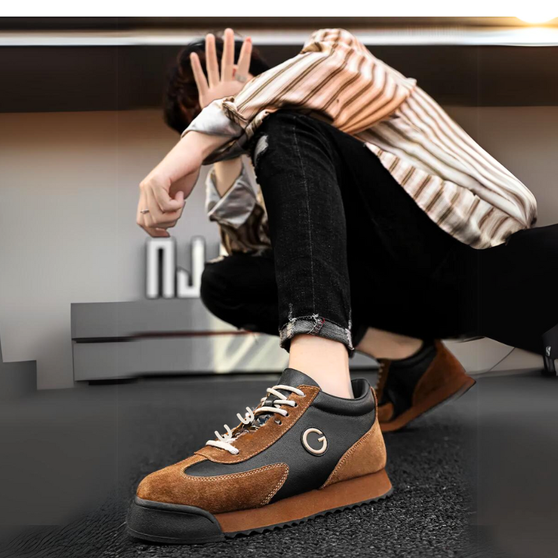 A person in a striped shirt and ripped black jeans sits on the ground, holding a hand up to partially cover their face. The focus is on their luxe leather suede sneakers with white laces and a G logo.