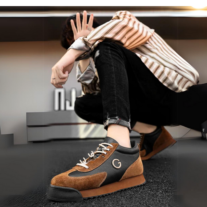 A person in a striped shirt and ripped black jeans sits on the ground, holding a hand up to partially cover their face. The focus is on their luxe leather suede sneakers with white laces and a G logo.