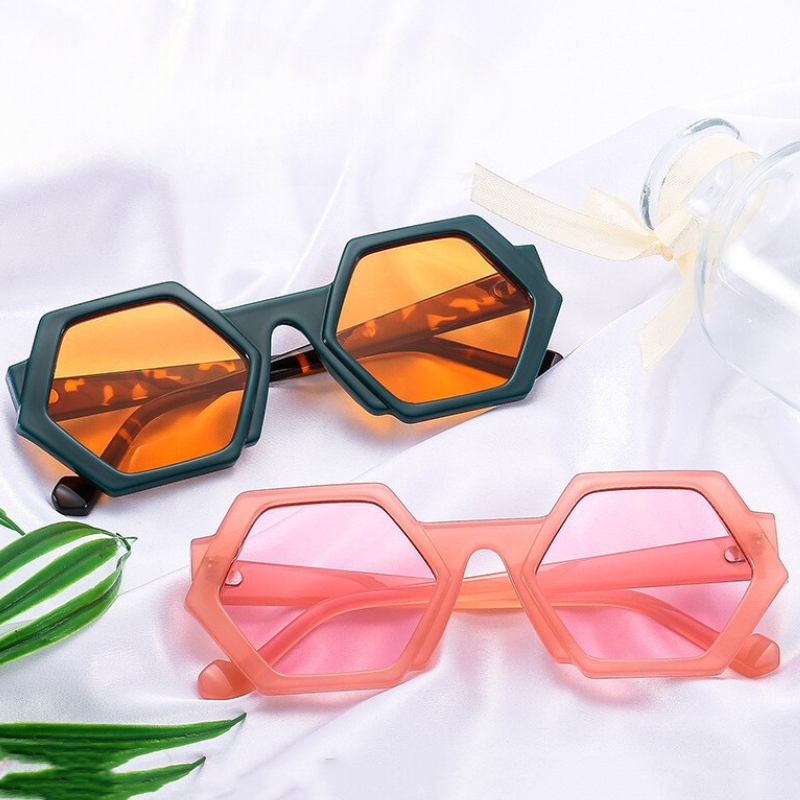 Two pairs of trend-forward hexagonal frame sunglasses rest on white fabric. One features dark green frames with orange lenses; the other, pink frames with light pink lenses. A clear bottle with a ribbon and green leaves completes the look.