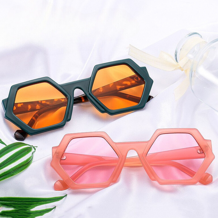 Two pairs of trend-forward hexagonal frame sunglasses rest on white fabric. One features dark green frames with orange lenses; the other, pink frames with light pink lenses. A clear bottle with a ribbon and green leaves completes the look.