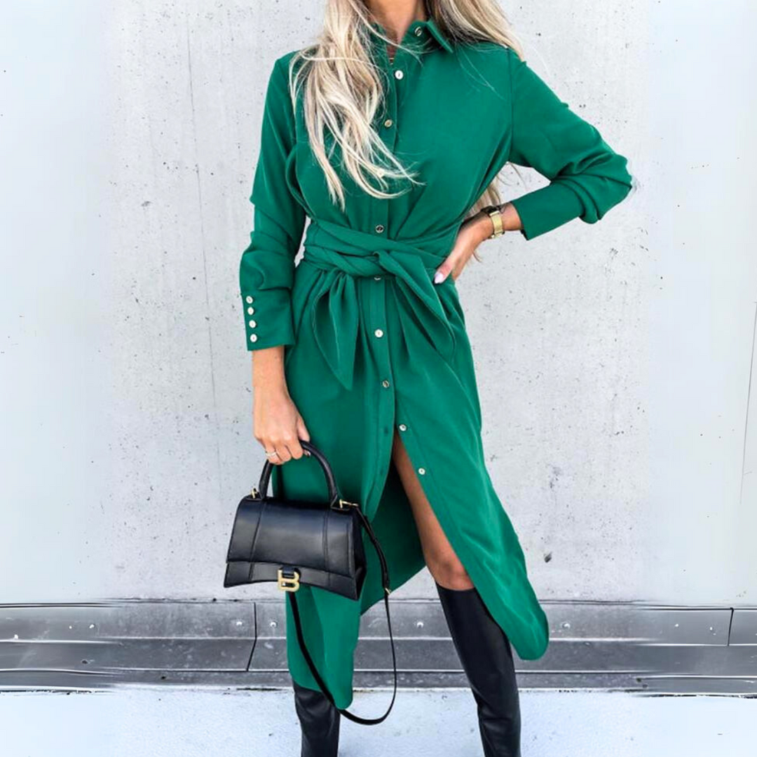 A woman in a long-sleeve wrap dress with a waist tie stands against a light gray wall. She pairs her green dress with black knee-high boots and a black handbag, blending streetwear style with high fashion flair. Her long blonde hair is loose.